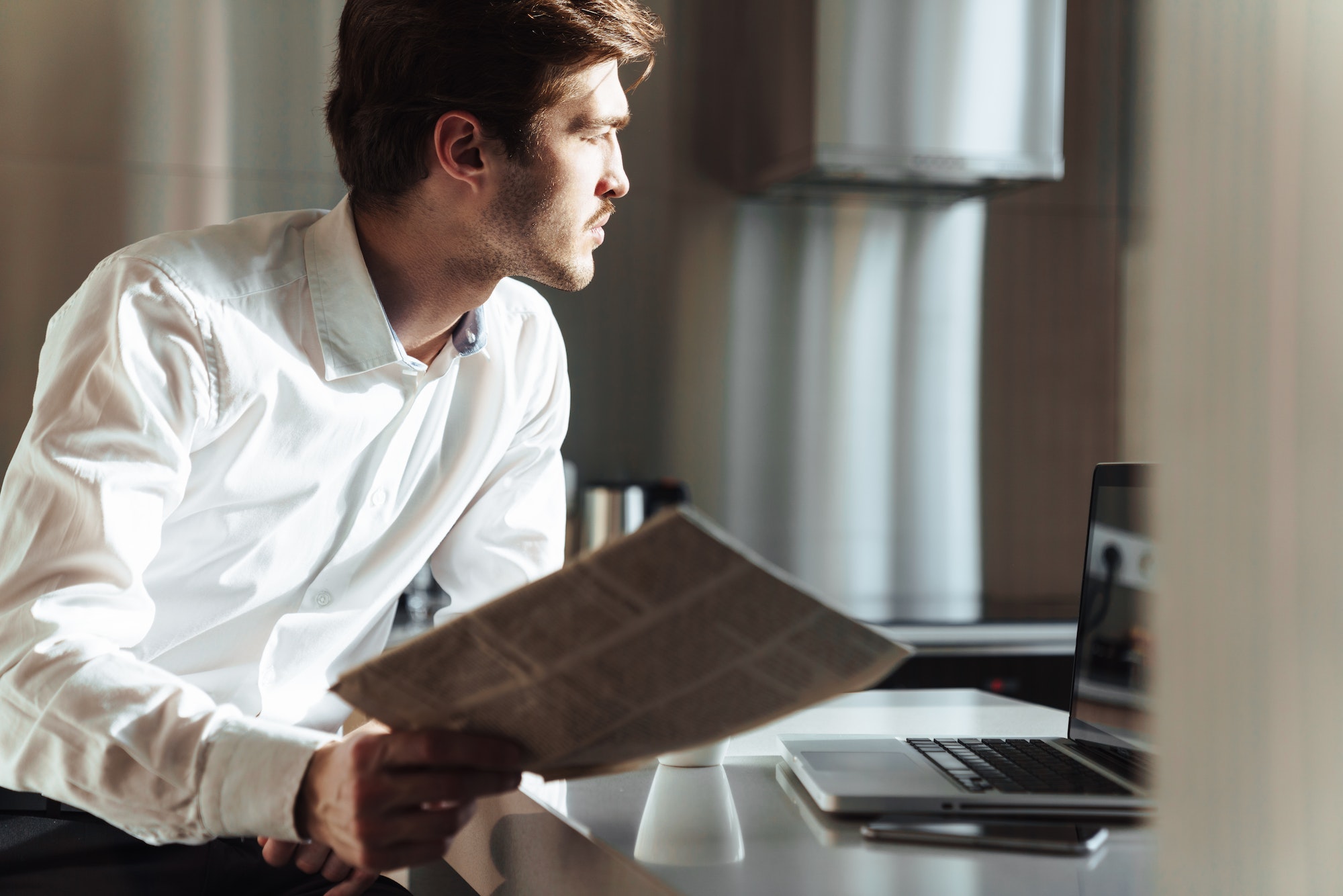 Photo of thinking businessman using laptop and reading newspaper